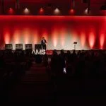 Dr Zack Ally presenting on stage at AMS25 in Malmö, speaking about facial sculpting to a full audience under red lighting