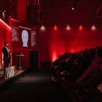 Dr Zack Ally presenting the LOOMIS Method to a large audience at AMS25, with anatomical visuals displayed on dual screens.